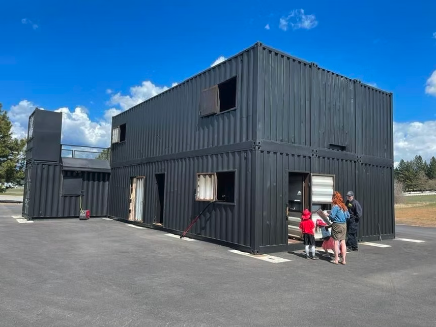 A black, two-story structure made from shipping containers with people standing outside on a sunny day.