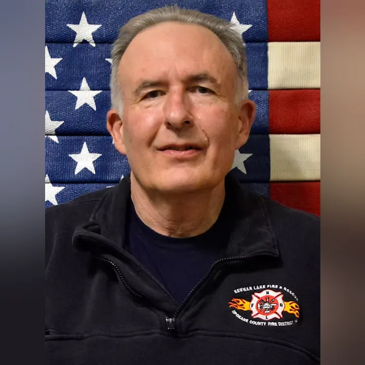A person in a dark jacket with fire department insignia stands in front of an American flag backdrop.
