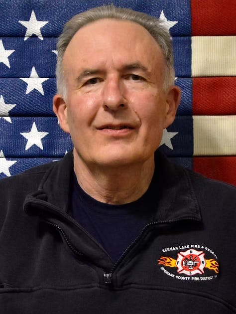 A person in a dark jacket with fire department insignia stands in front of an American flag backdrop.
