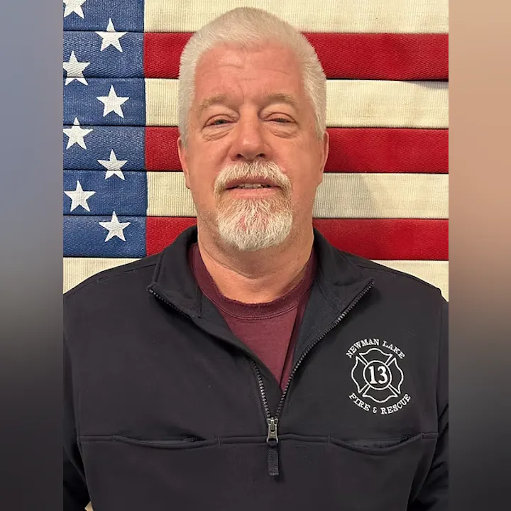 A man with white hair and beard is standing in front of an American flag, wearing a Newman Lake Fire & Rescue jacket.