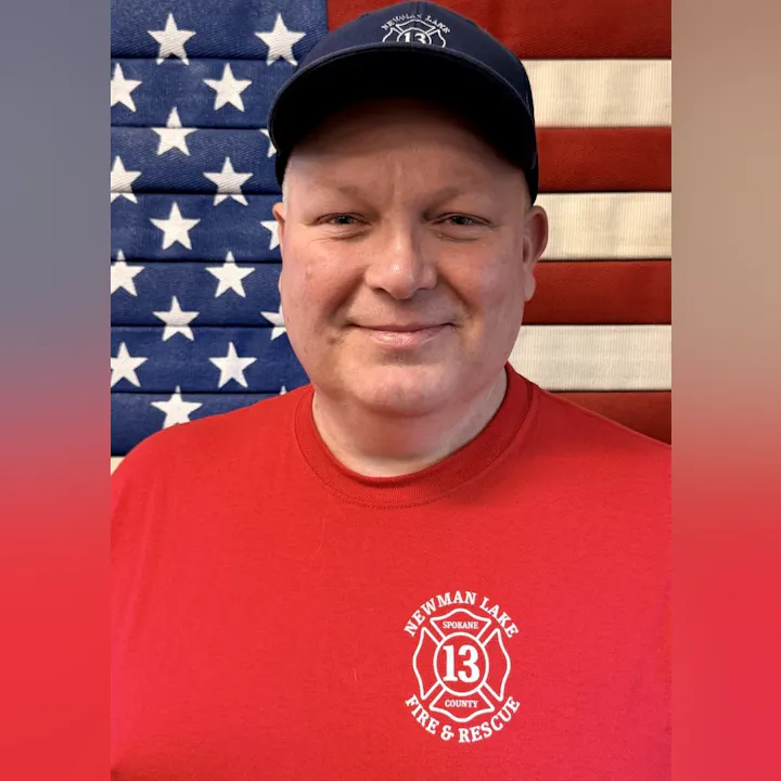 A smiling person in a red shirt with a fire department logo, posed in front of an American flag backdrop.
