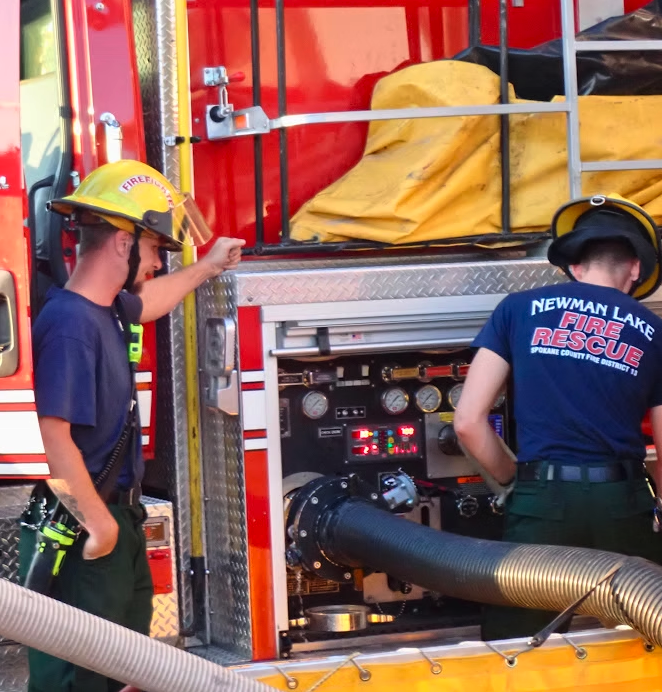 Two firefighters beside a fire truck operating equipment with hoses attached.