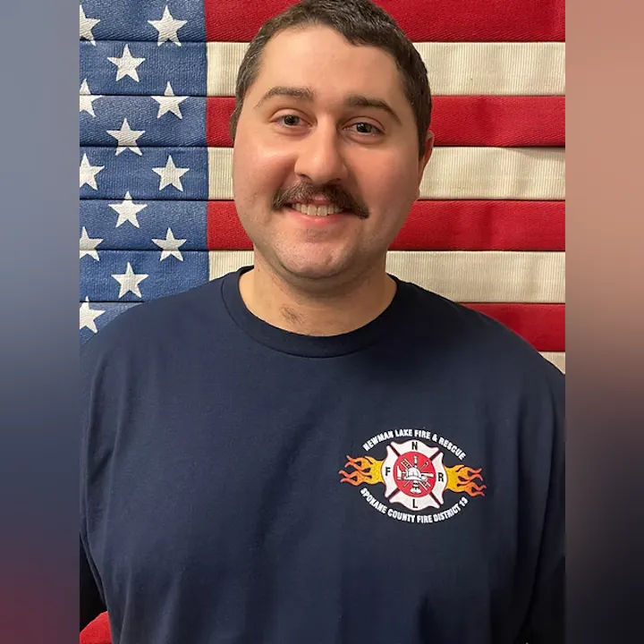 A person in a fire department shirt stands in front of an American flag backdrop.