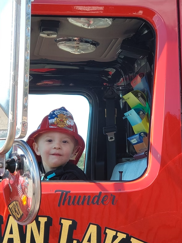 A child wearing a red fire helmet is sitting inside a fire truck with "Thunder" written on the side.