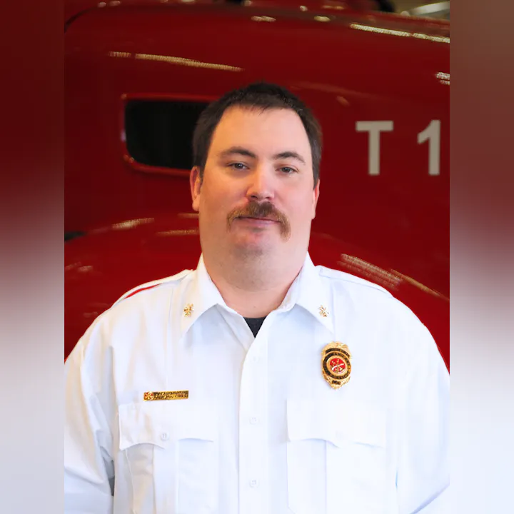A man in a white uniform with a badge stands in front of a red fire truck, smiling for the camera.