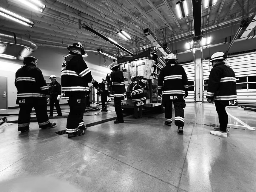 Firefighters in a station with a fire truck, wearing uniforms and helmets, appear to be in a training or preparation scenario.