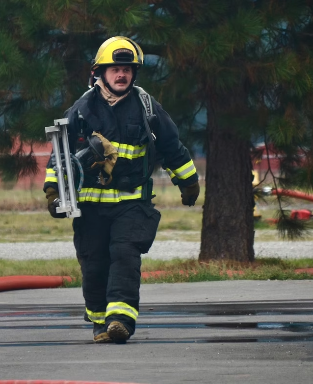 A firefighter in gear carrying a metal ladder, walking on a wet pavement with trees in the background.