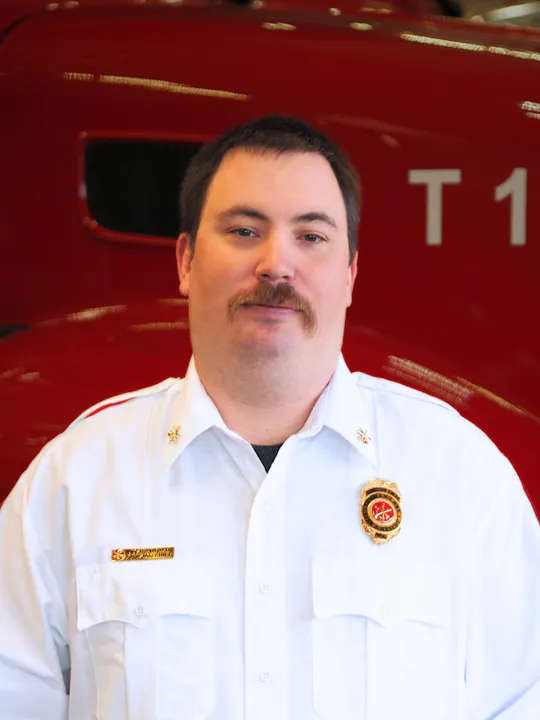 The image shows a man in a white uniform with a badge, standing in front of a red vehicle.
