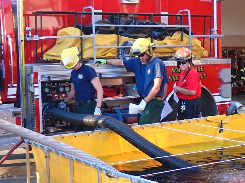 Firefighters operate equipment from a fire truck with hoses and a portable tank nearby.