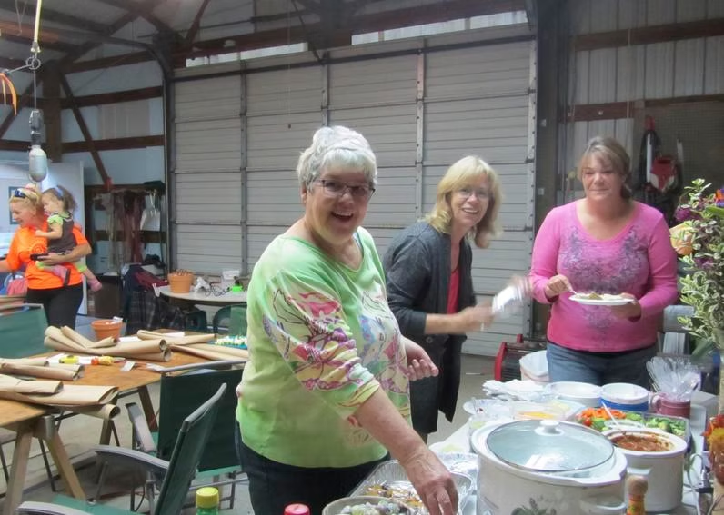 People serving food at a gathering in a garage with tables and dishes.