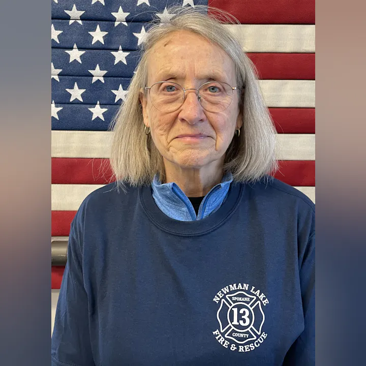 An older woman stands in front of an American flag, wearing a fire department shirt from Newman Lake Fire & Rescue.