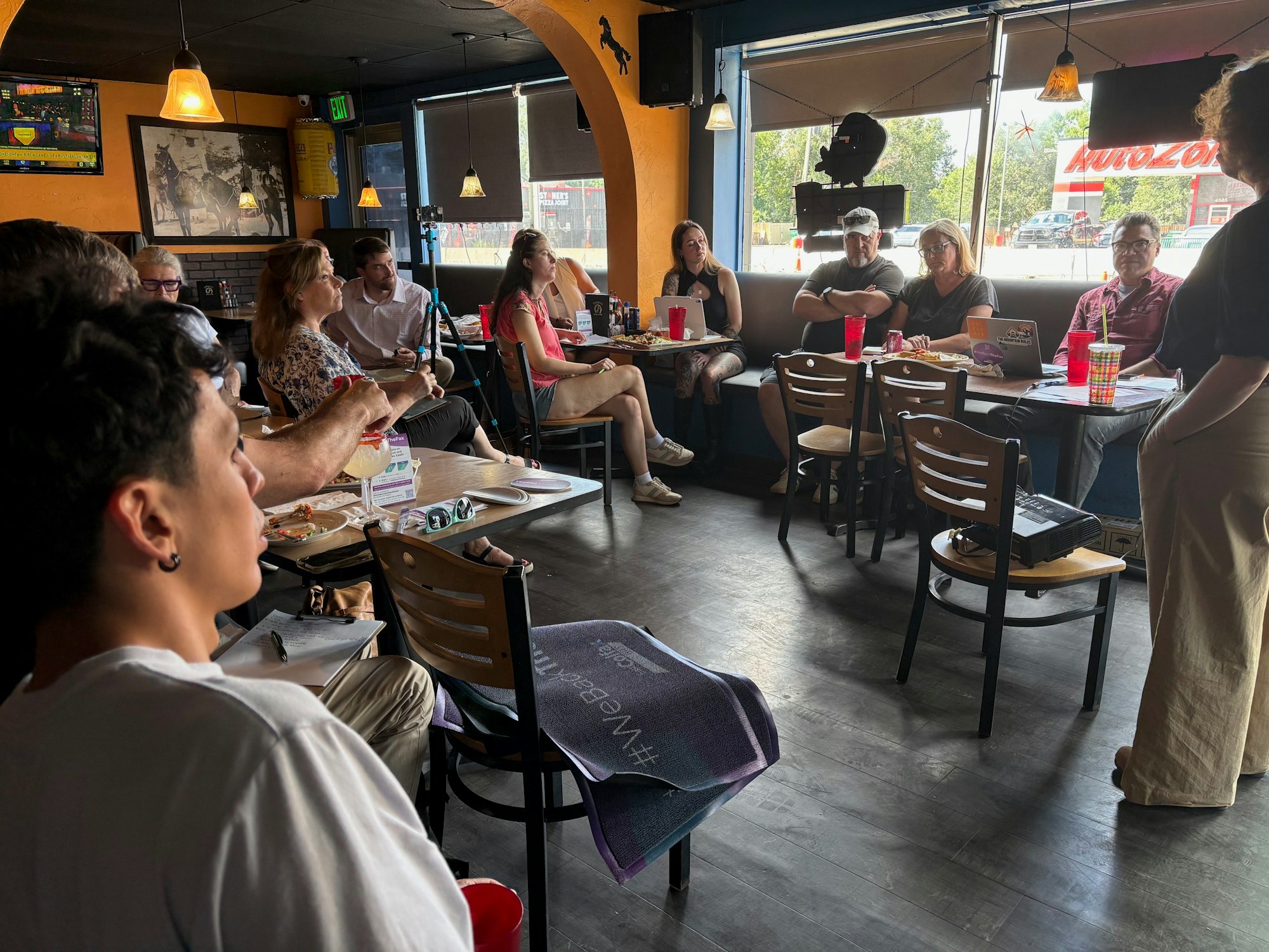 A group of people in a cafe, engaging in a discussion, with food and drinks on tables, under warm lighting.