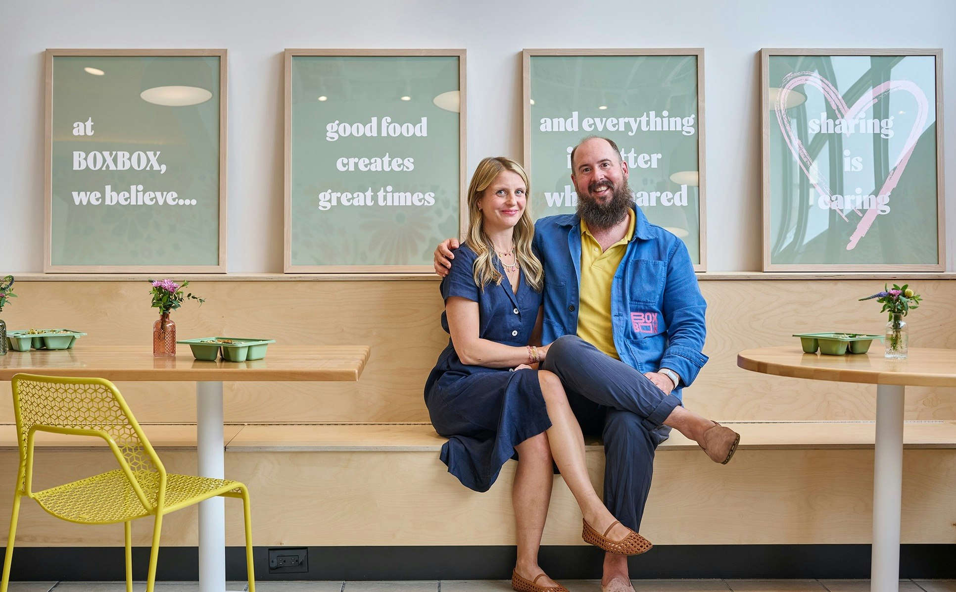 A couple sitting in a restaurant booth, smiling; framed quotes behind them promoting good food and sharing.