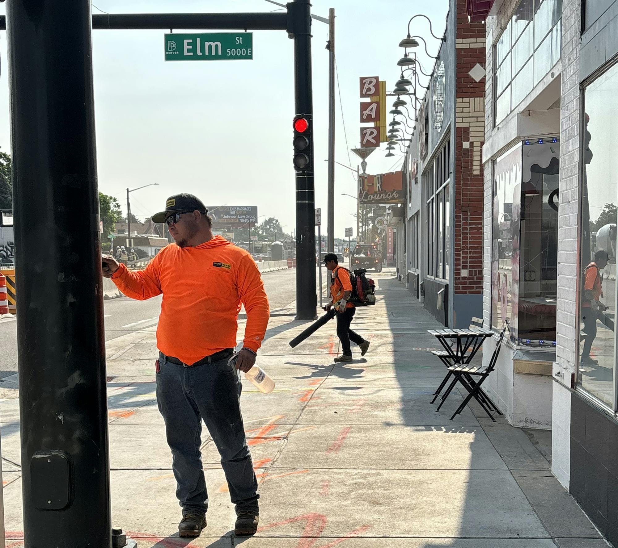 Two workers in orange shirts on Elm Street; one checking a device, the other using a leaf blower.