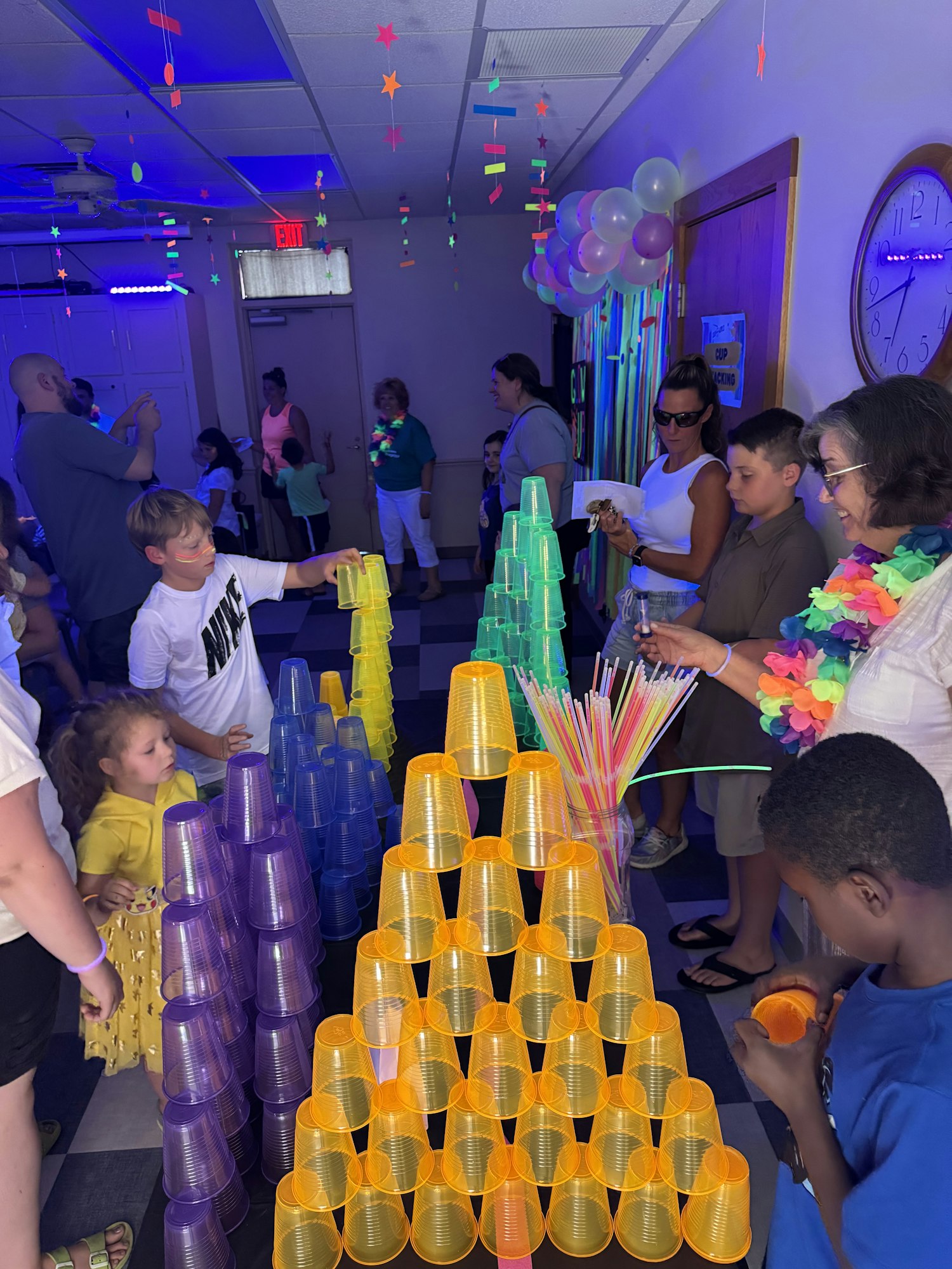 People are stacking colorful cups under blacklight, with glow-in-the-dark decorations and balloons in the background.