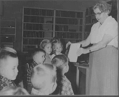 A woman shows a book to a group of children in a library setting.