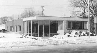 A single-story building with large windows, surrounded by snow, trees, and a road in the foreground.