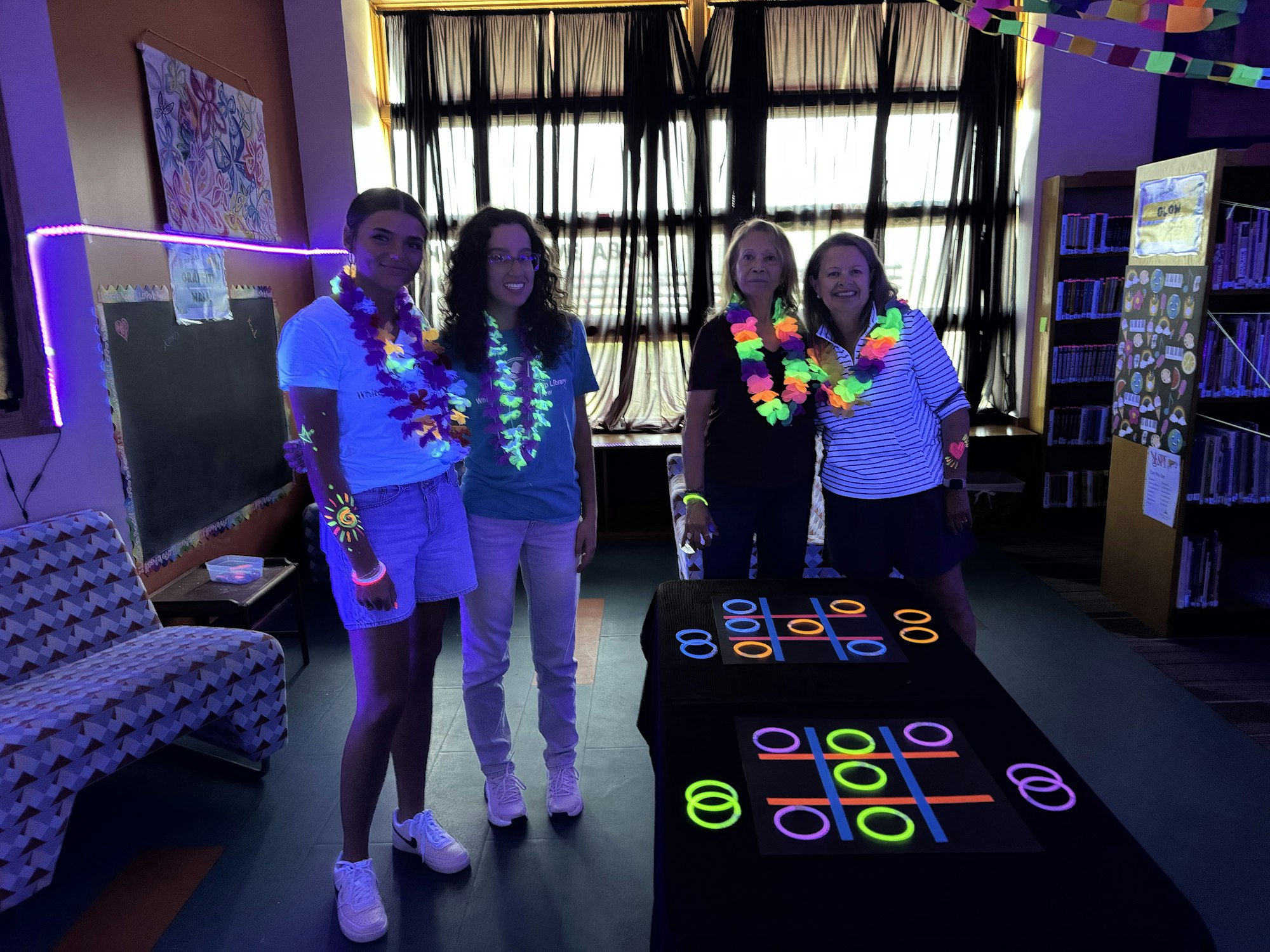 Four people under blacklight with glowing accessories, next to a tic-tac-toe game with neon circles and crosses.