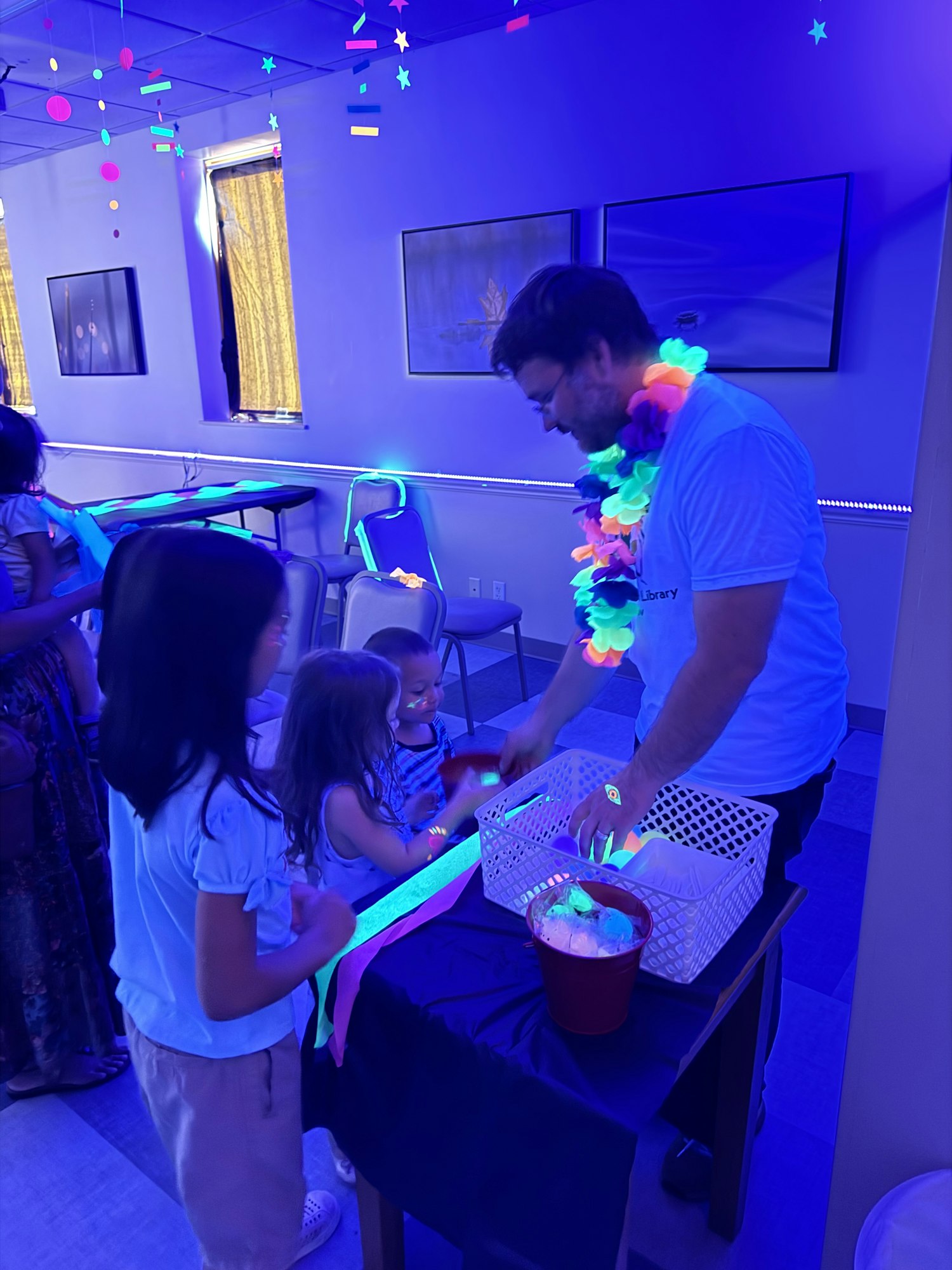 A person with a colorful lei and kids are enjoying a glow-in-the-dark activity in a neon-lit room.