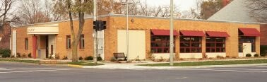 A small brick building with red awnings, situated on a street corner with trees and a bench nearby.
