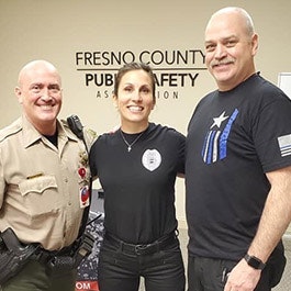 Three people smiling, two in uniform, one with a "Thin Blue Line" shirt, in front of a "Fresno County Public Safety Association" sign.
