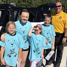 A group of kids in matching blue shirts with an adult standing by a vehicle, all smiling.