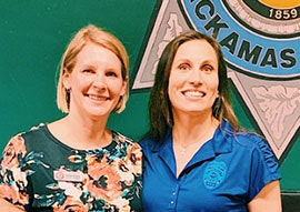 Two women smiling in front of a sign that reads "Clackamas 1859."