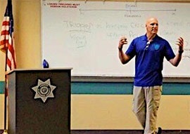A person presenting in a classroom with a whiteboard and a U.S. flag.