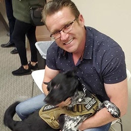 A man smiling, holding a happy dog wearing a harness in his lap.