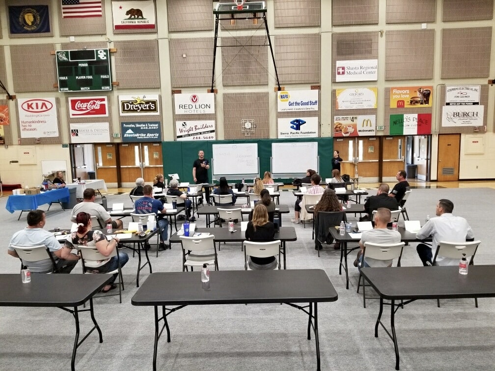 A training or workshop session in a gymnasium with participants seated at tables and presenters at the front.