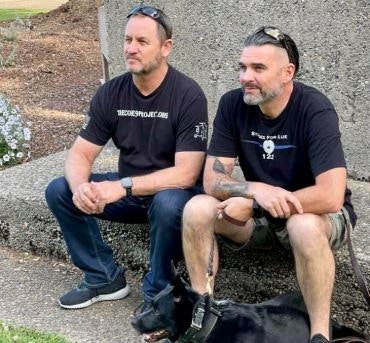 Two men squatting next to a dog, wearing black shirts with text, outdoors.