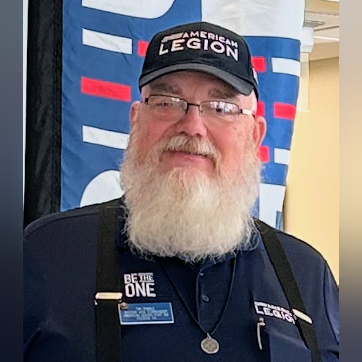 A bearded man in an "American Legion" hat and shirt stands in front of a banner.