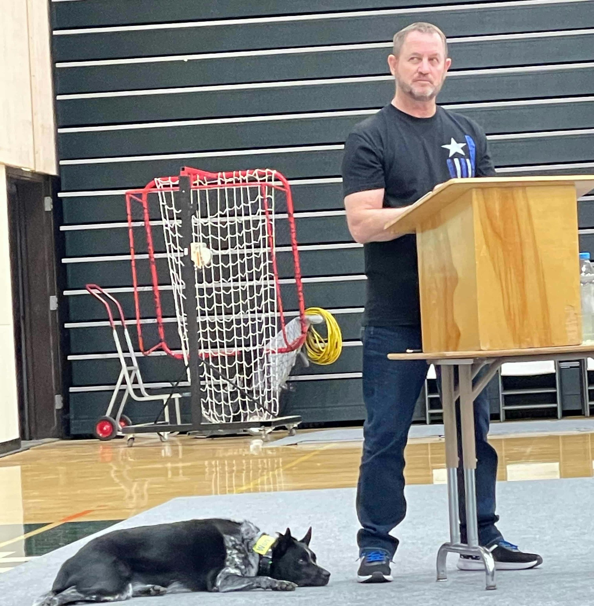 A man standing at a podium with a dog lying on the ground in a gym, next to a hockey net and chairs.