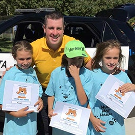 Three people with certificates, standing by a car on a sunny day. Two are children, one adult, all smiling.