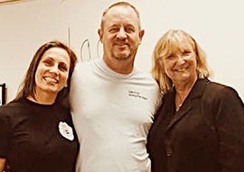 Three people smiling for a photo, two women flanking a man, in a room with a white background.
