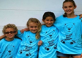 Four children smiling, wearing blue T-shirts with a polar bear print and the message "HUGS NOT HARM."
