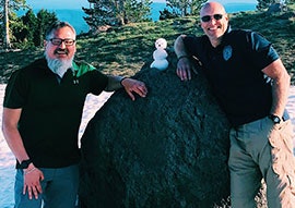 Two men smiling next to a small snowman on a rock, with trees and clear blue sky in the background.