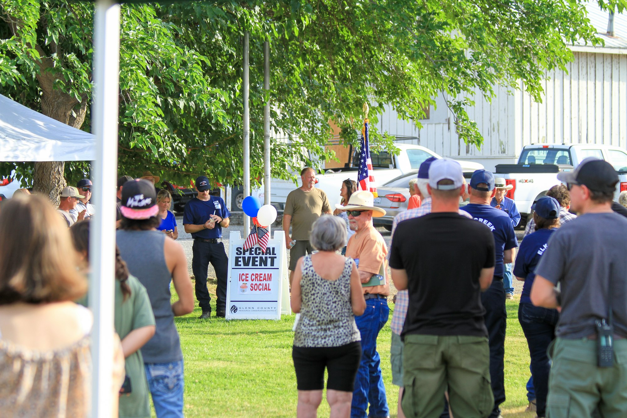 Outdoor community event with people, an "Ice Cream Social" sign, balloons, and an American flag.