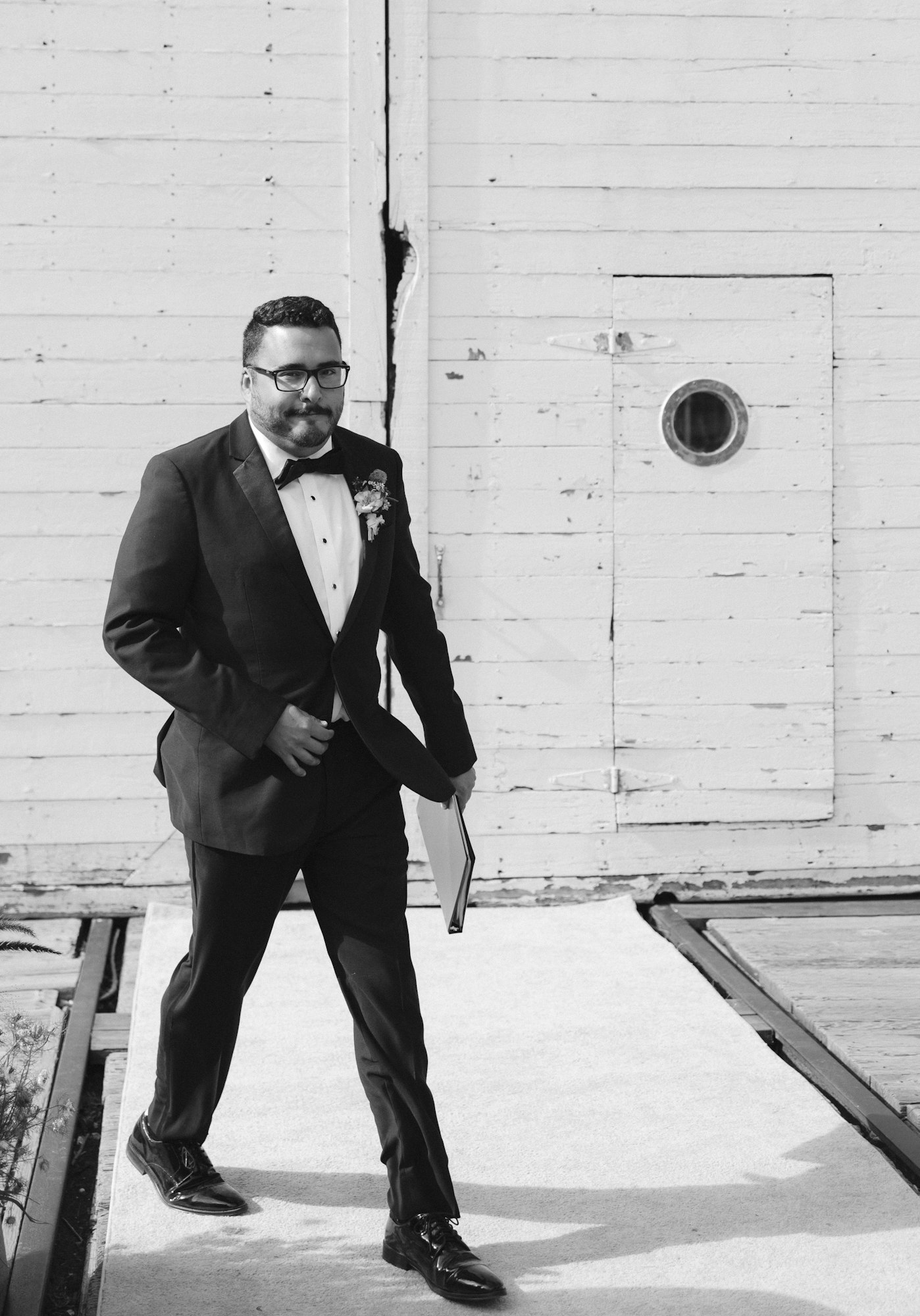 Man in a suit walking on a path, holding a book, with a wooden wall background.