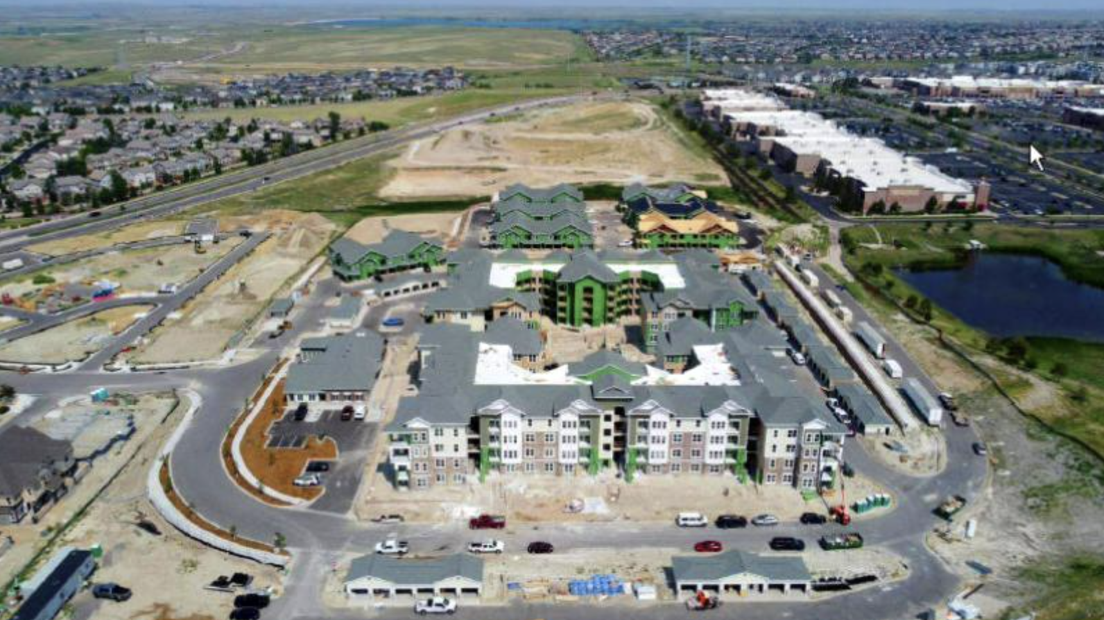 Aerial view of a suburban development with apartment buildings under construction, roads, and a pond.