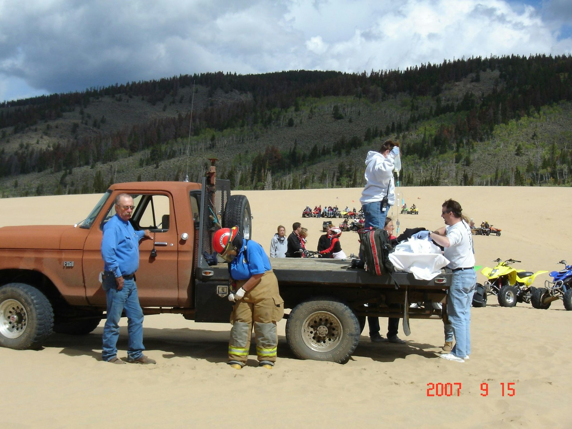 People by a truck on dunes with ATVs in the background under a cloudy sky. Date stamp: 2007-09-15.