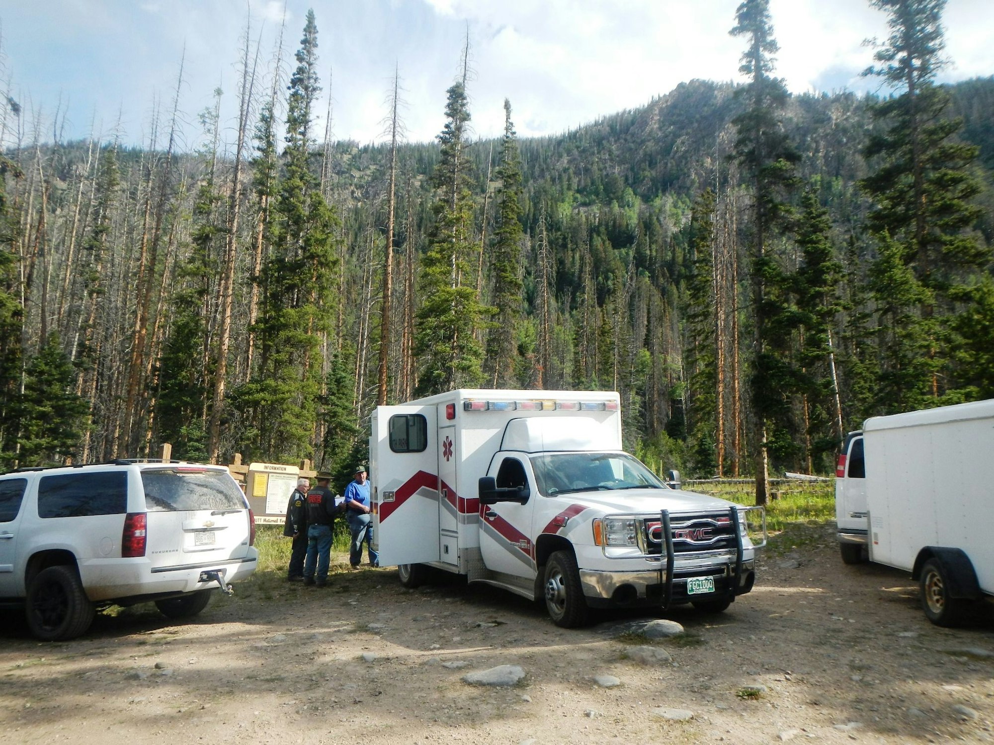 Ambulance and vehicles in a forested area with people standing by.