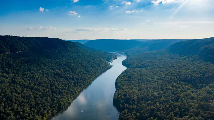 A scenic view of a river winding through lush green hills under a blue sky with scattered clouds.