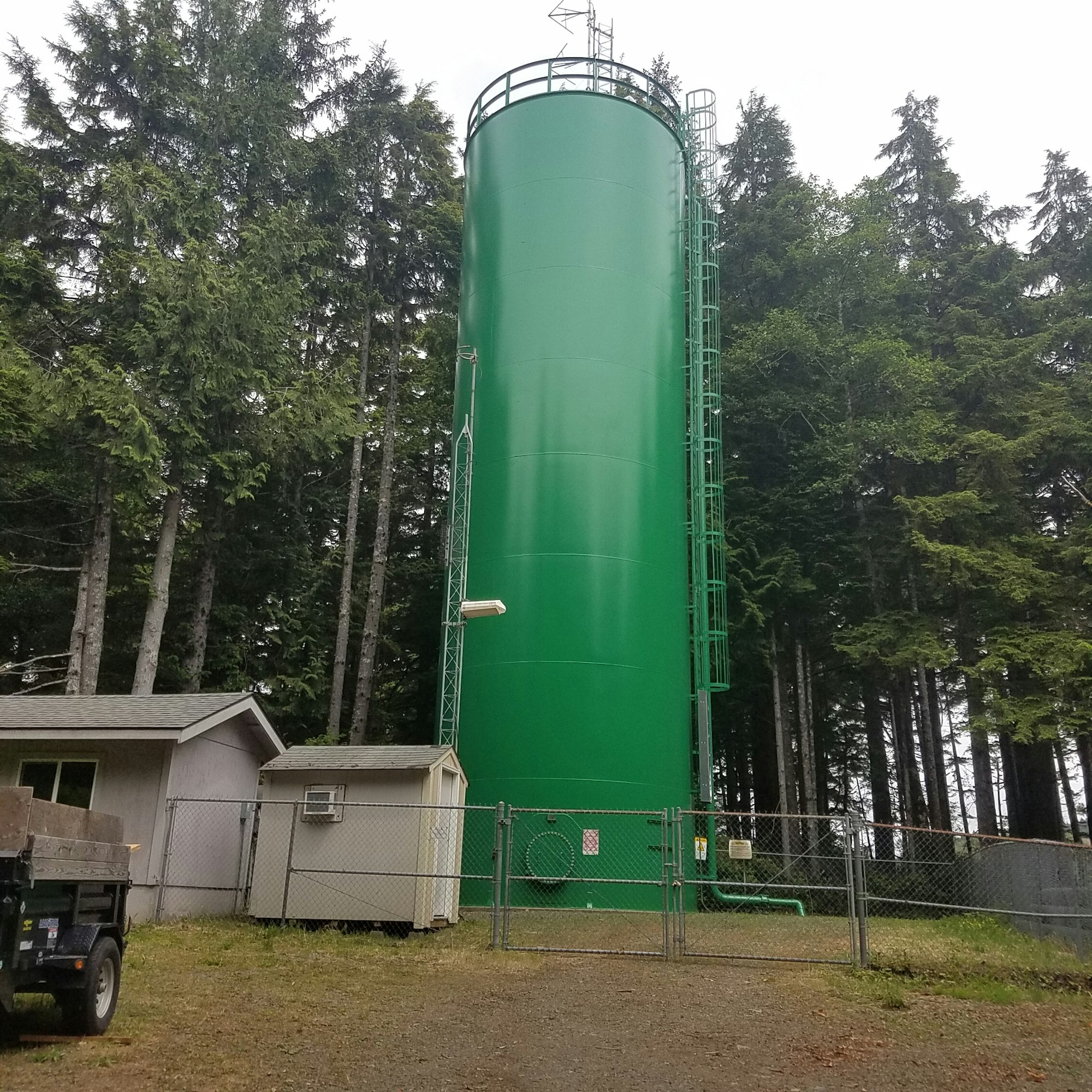 A large green storage tank with a fence around it, surrounded by trees. Image is rotated 90 degrees to the right.