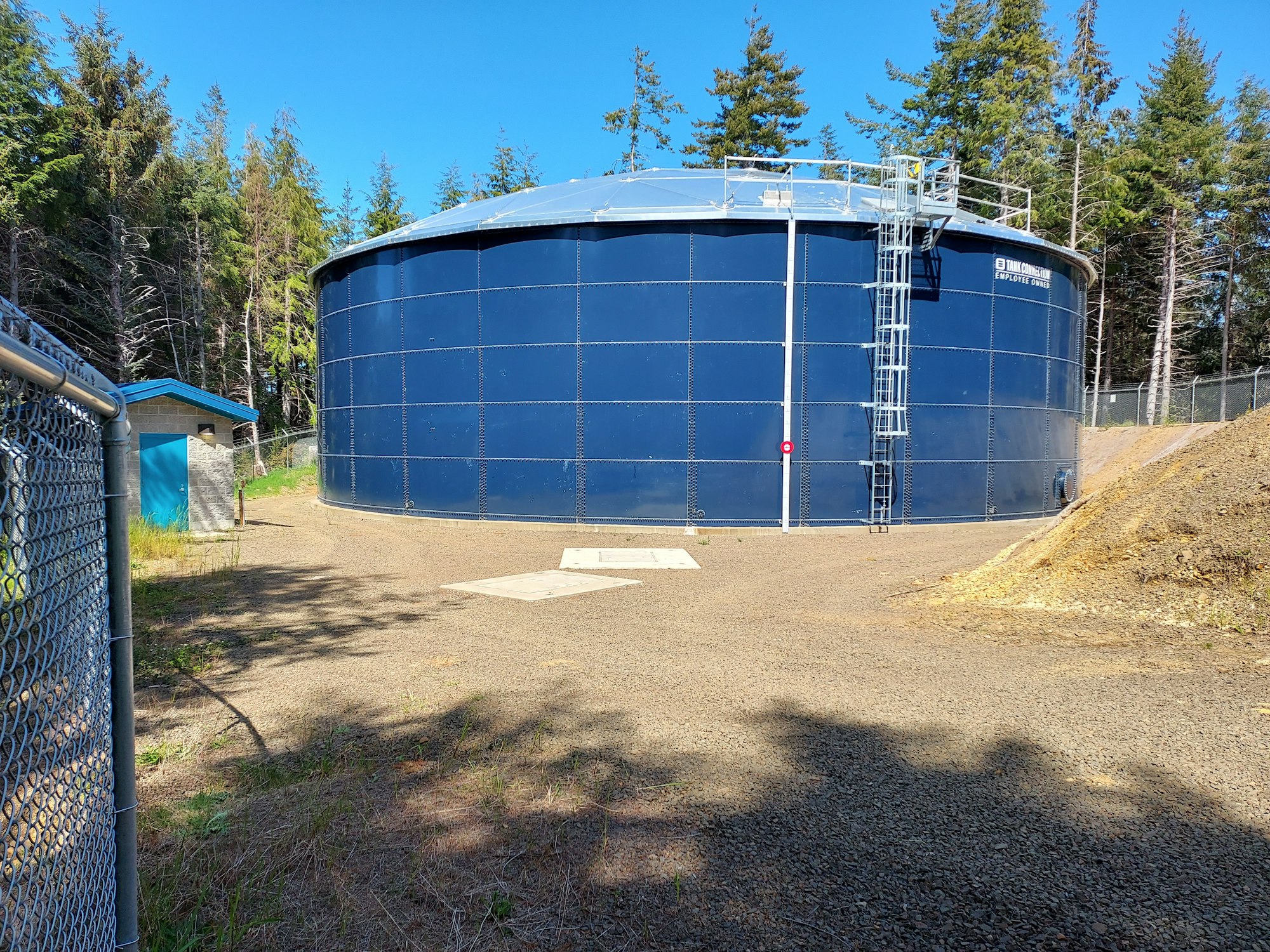 A large blue water storage tank with surrounding fencing, set against a wooded area under a blue sky.