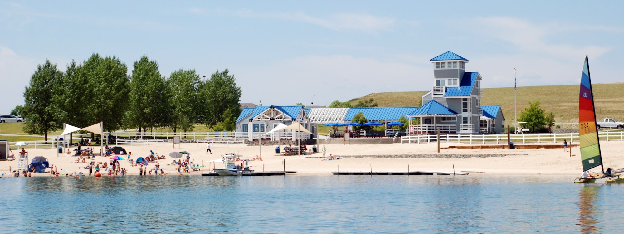 A lakeside beach scene with people, a pier, boats, and buildings with blue roofs under a clear sky.