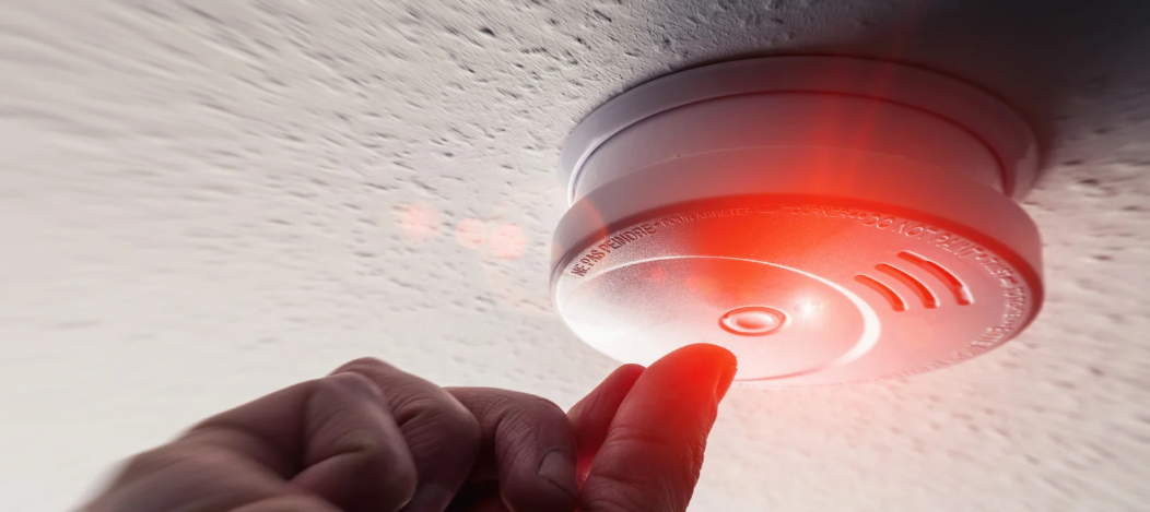 A hand pressing a test button on a smoke detector with a red light.
