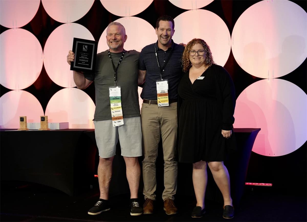 Three people are smiling for a photo on stage; one is holding an award. There are also three trophies on a table behind them.