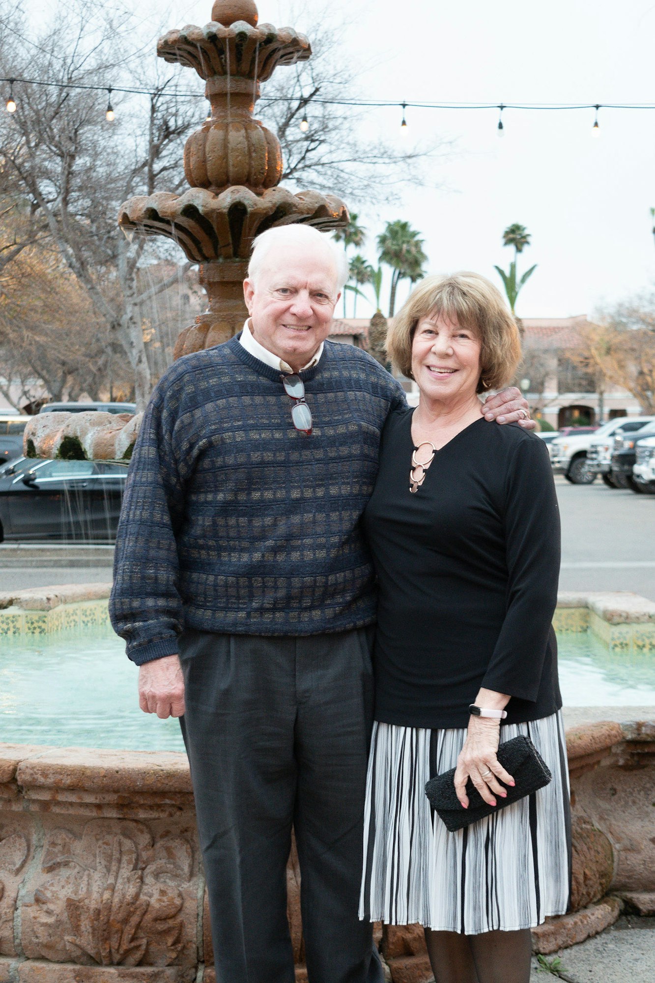 A smiling couple posing before a fountain with string lights and palm trees in the background.