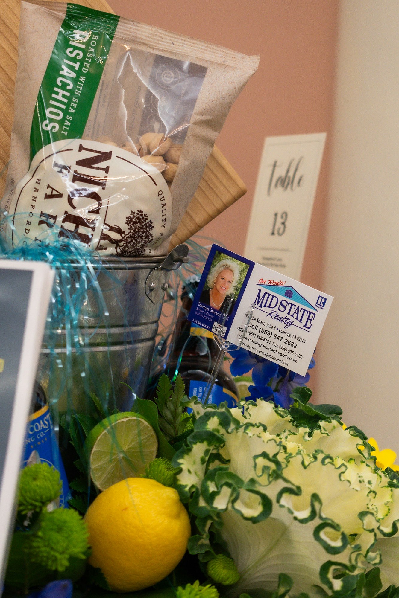 Gift basket with snacks, drinks, flowers, lemon, business card, and "Table 13" sign.